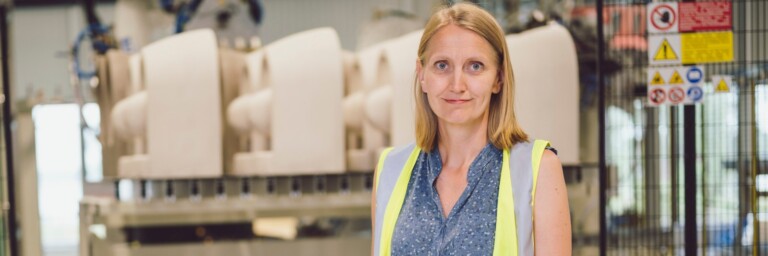 Eine Frau mit einem blauen Oberteil und einer gelben Warnweste steht vor industriellen Maschinen in einer Fabrik.