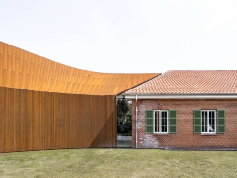 A modern architectural structure with a curved wooden facade adjacent to a traditional brick house with green shuttered windows.