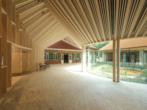 Interior view of a modern building with a high, sloped wooden ceiling, large windows, and an open layout, featuring a courtyard garden.