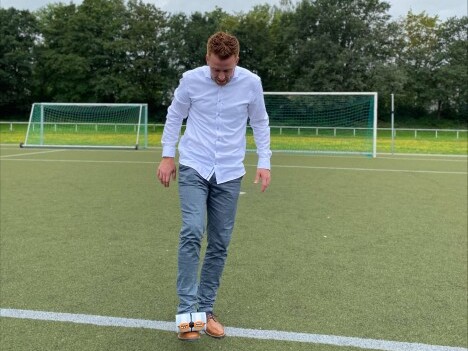 Ein Mann in einem weissen Hemd und Jeans steht auf einem Fussballplatz und balanciert etwas auf seinem Fuss.