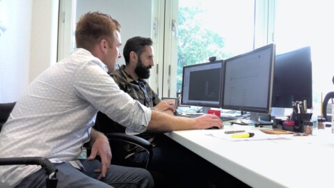 Zwei Männer sitzen an einem Schreibtisch, schauen auf Bildschirme und arbeiten an einem Computerprojekt.