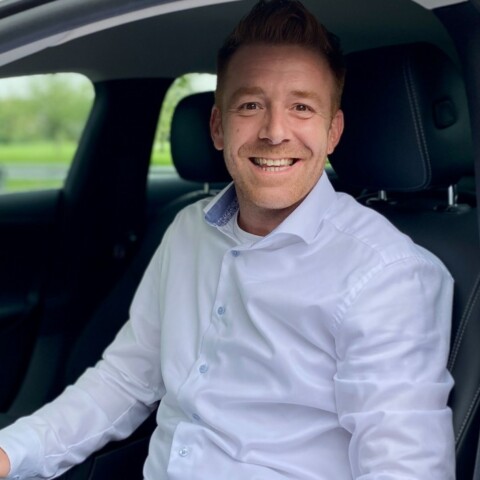 Ein lächelnder Mann in einem weissen Hemd sitzt in einem Auto und schaut zur Kamera.