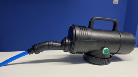 A black plastic device with a handle, featuring a hose and a green button, set against a blue background.