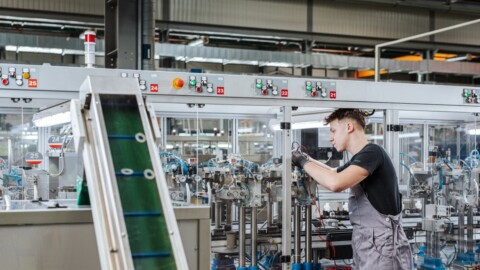 A worker wearing gloves adjusts machinery in a manufacturing facility with industrial equipment visible in the background.