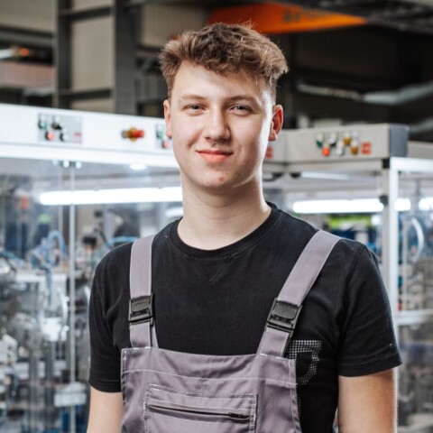 A young man in gray overalls stands smiling in a factory setting with machinery in the background.