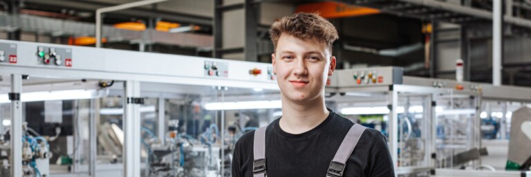 A young man in gray overalls stands smiling in a factory setting with machinery in the background.