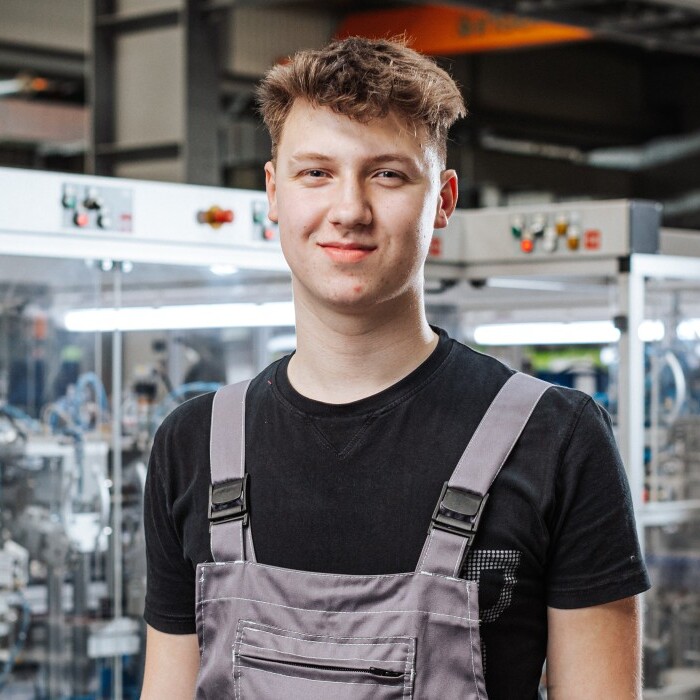 A young man in gray overalls stands smiling in a factory setting with machinery in the background.