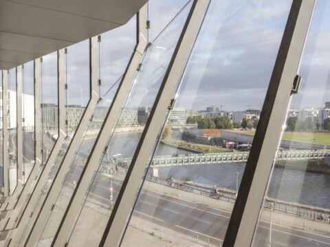 A modern architectural interior with large angled windows overlooking a cityscape, river, and bridge under cloudy skies.