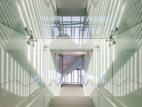 A modern staircase with glass walls leading to a bright area with large windows.