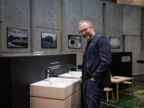 Ein Mann in Brille steht lächelnd neben einem modernen Waschbecken in einer Ausstellungsumgebung mit Fotografien an der Wand.