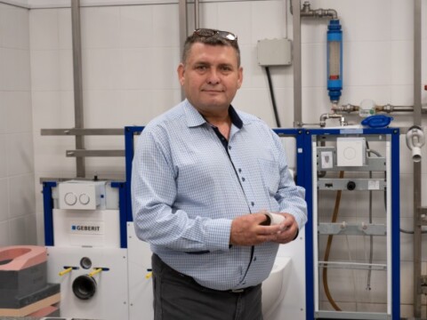 Ein Mann in einem hellblauen Hemd steht in einem technischen Raum mit Geräten im Hintergrund.