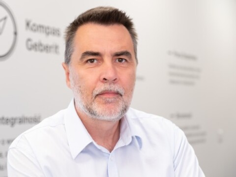 Ein Mann mit grauem Haar und Bart sitzt vor einer hellen Wand mit Text.