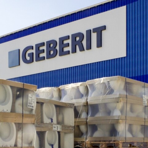 Lagerbereich mit Paletten von Sanitärprodukten vor einer blauen Geberit-Fabrikfassade.