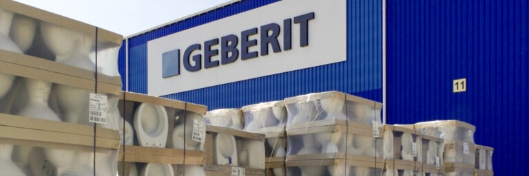 Lagerbereich mit Paletten von Sanitärprodukten vor einer blauen Geberit-Fabrikfassade.