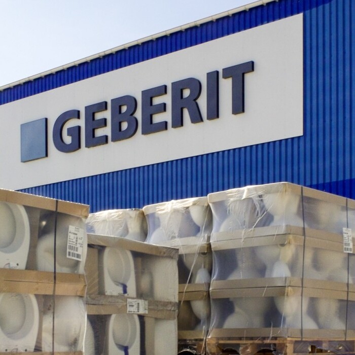 Lagerbereich mit Paletten von Sanitärprodukten vor einer blauen Geberit-Fabrikfassade.