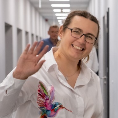 A woman with glasses wearing a white shirt with a colourful bird design waves, smiling in a corridor.