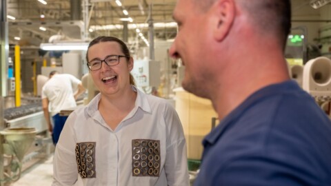 A woman with glasses and a white shirt with unique patterns laughs while engaging in conversation with a man in a workshop setting.