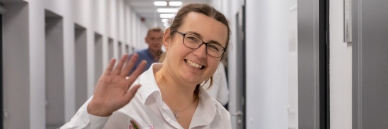 A smiling woman wearing glasses and a white shirt waves while standing in a corridor.