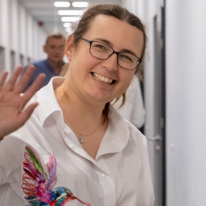 A smiling woman wearing glasses and a white shirt waves while standing in a corridor.