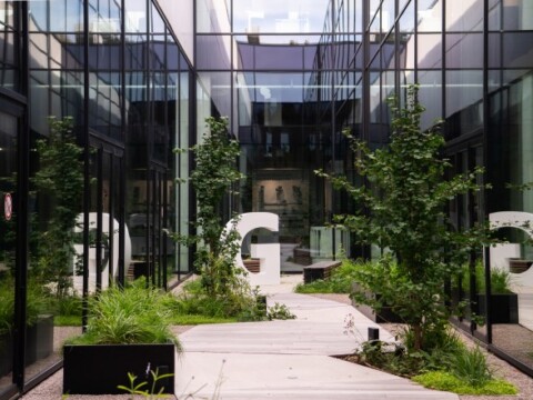 A modern courtyard with glass walls, greenery, and a large white letter "G", featuring a wooden walkway.