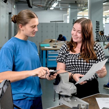 Two individuals collaborate in a workshop, one holding a tool while the other holds a sheet of paper, surrounded by equipment and work surfaces.