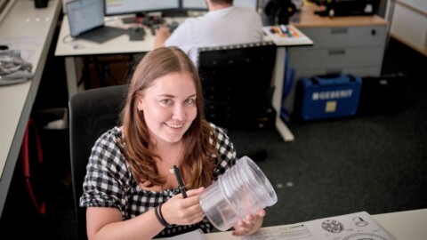A young woman with long hair, wearing a black and white checkerd shirt, smiling and holding a clear tube.