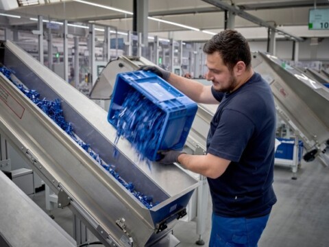 A worker in a manufacturing facility is pouring blue plastic items from a container onto a conveyor belt.