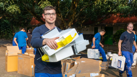 A group of individuals in blue uniforms carrying boxes and equipment outdoors, with greenery and buildings in the background.