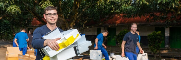 A group of individuals in blue uniforms carrying boxes and equipment outdoors, with greenery and buildings in the background.