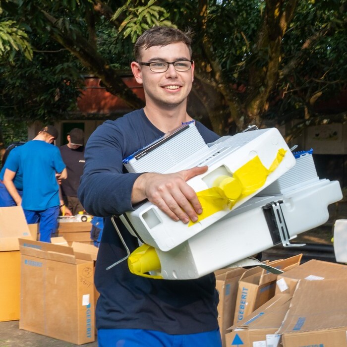 A group of individuals in blue uniforms carrying boxes and equipment outdoors, with greenery and buildings in the background.