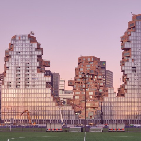Modern architectural buildings with a fragmented design, set against a pastel sunset sky, located next to a green sports field.