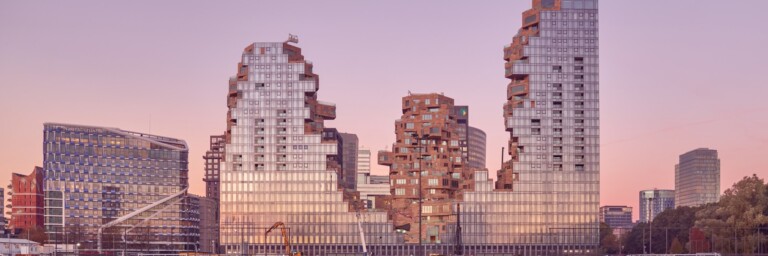 Modern architectural buildings with a fragmented design, set against a pastel sunset sky, located next to a green sports field.