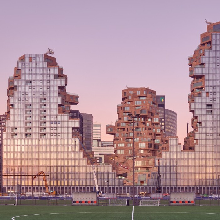 Modern architectural buildings with a fragmented design, set against a pastel sunset sky, located next to a green sports field.