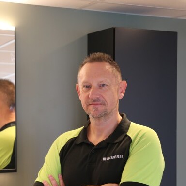 Mann in einem Schwarz-Grünen Poloshirt mit verschränkten Armen, steht vor einem Spiegel in einem modernen Innenraum.