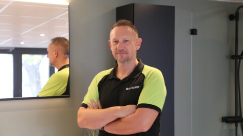 Ein Mann steht mit verschränkten Armen vor einem Spiegel in einem modernen Innenraum. Er trägt ein schwarzes und grünes Polohemd.