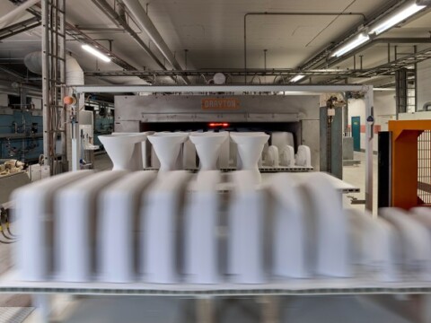 Ein keramisches Fertigungswerk mit weissen Formen, die auf einem Förderband vorbeigeführt werden, und einem Brennofen im Hintergrund.