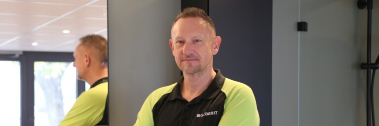 Ein Mann steht mit verschränkten Armen vor einem Spiegel in einem modernen Innenraum. Er trägt ein schwarzes und grünes Polohemd.