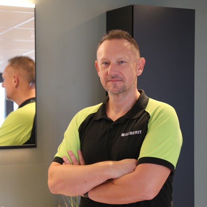 Ein Mann steht mit verschränkten Armen vor einem Spiegel in einem modernen Innenraum. Er trägt ein schwarzes und grünes Polohemd.