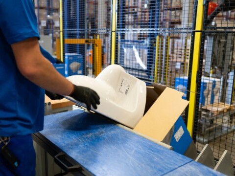 Ein Mitarbeiter in blauer Kleidung und Handschuhen schiebt ein weisses Toilettenbecken in einen Karton in einer Lagerumgebung.