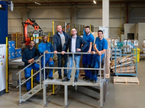 Sechs Personen in blauen Arbeitskleidung stehen auf einer Metallplattform in einer industriellen Umgebung mit Maschinen und Regalen im Hintergrund.