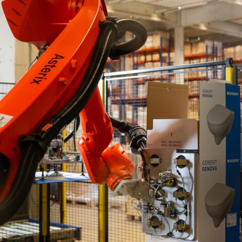 Ein roter Industrieroboterarm arbeitet in einem Lager mit verschiedenen Produkten im Hintergrund.