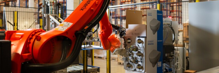 Ein roter Industrieroboterarm arbeitet in einem Lager mit verschiedenen Produkten im Hintergrund.