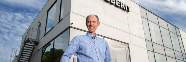 Mann in einem blauen Hemd steht vor einem modernen Firmengebäude mit dem Schriftzug "Geberit".