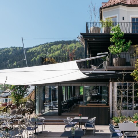 Moderne Terrasse eines Restaurants mit Markisen, Tischen und Stühlen, umgeben von Bepflanzung und Blick auf die Berge.