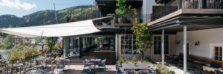 Moderne Terrasse eines Restaurants mit Markisen, Tischen und Stühlen, umgeben von Bepflanzung und Blick auf die Berge.