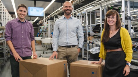 Drei Personen stehen in einer industriellen Umgebung neben grossen Kartons, lächeln und posieren freundlich.