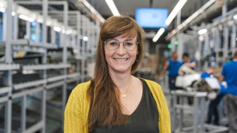 Eine lächelnde Frau mit langen braunen Haaren und Brille steht in einem modernen Arbeitsumfeld.