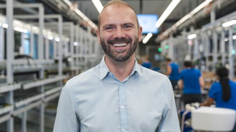 Mann mit Bart und lächelndem Gesicht, der in einer modernen Produktions- oder Lagerumgebung steht.