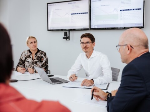 A group of professionals in a meeting, discussing information displayed on two screens, with a laptop and notepads on the table.