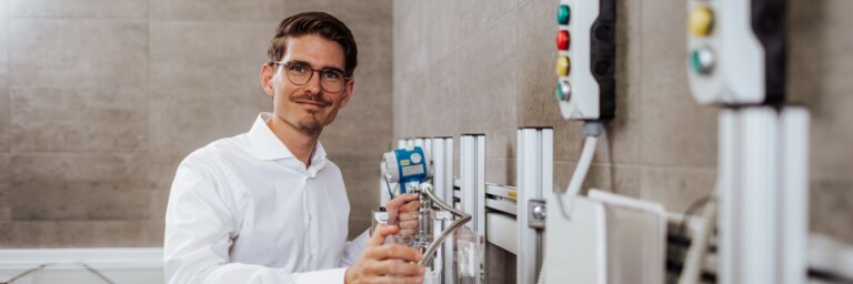 A man in a white shirt holding a hose and smiling, standing in front of control panels with buttons.